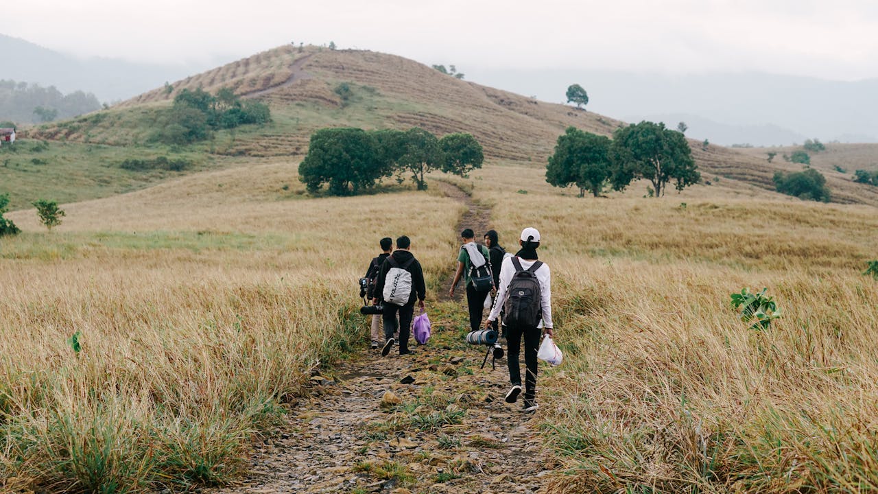 Back view of a group hiking through a rural field towards a hill under an overcast sky.